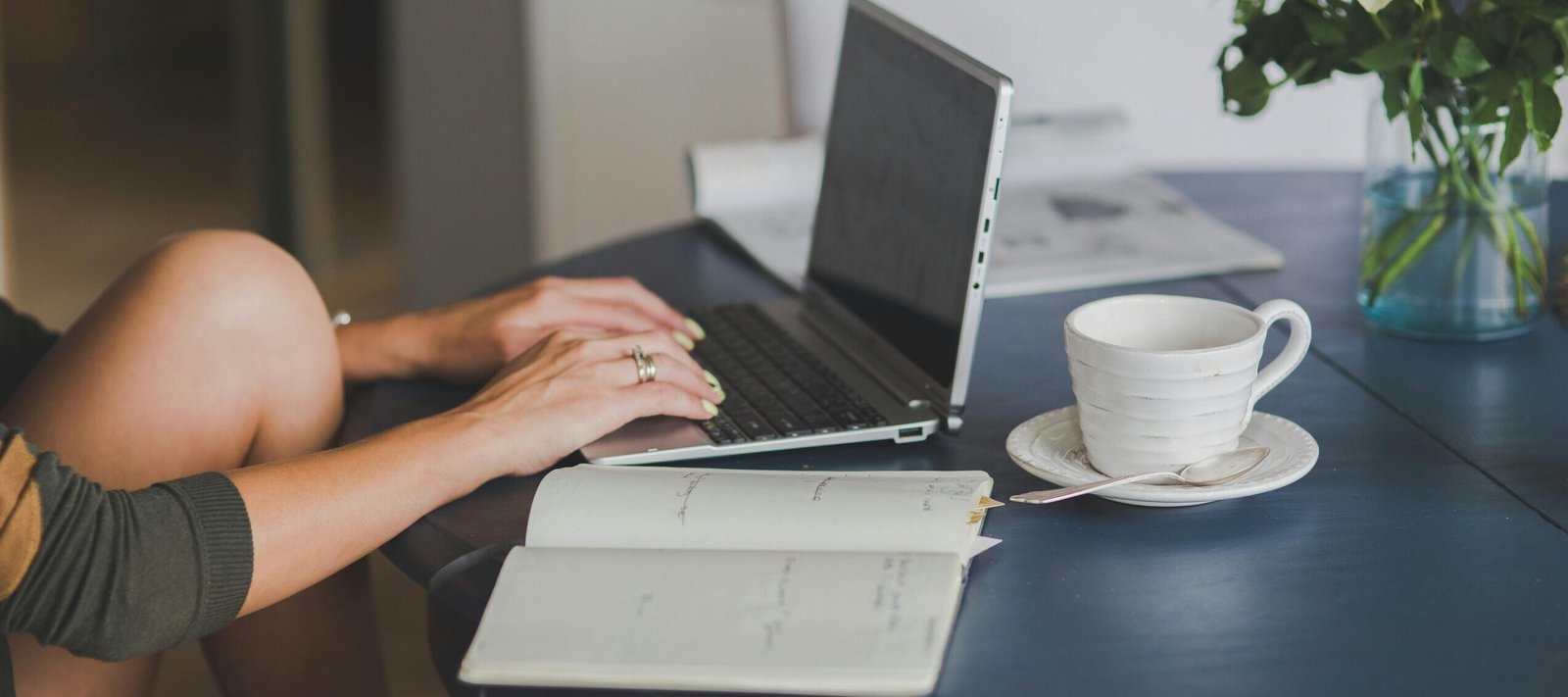 Female freelancer using laptop with coffee at home office desk, surrounded by roses and a planner.